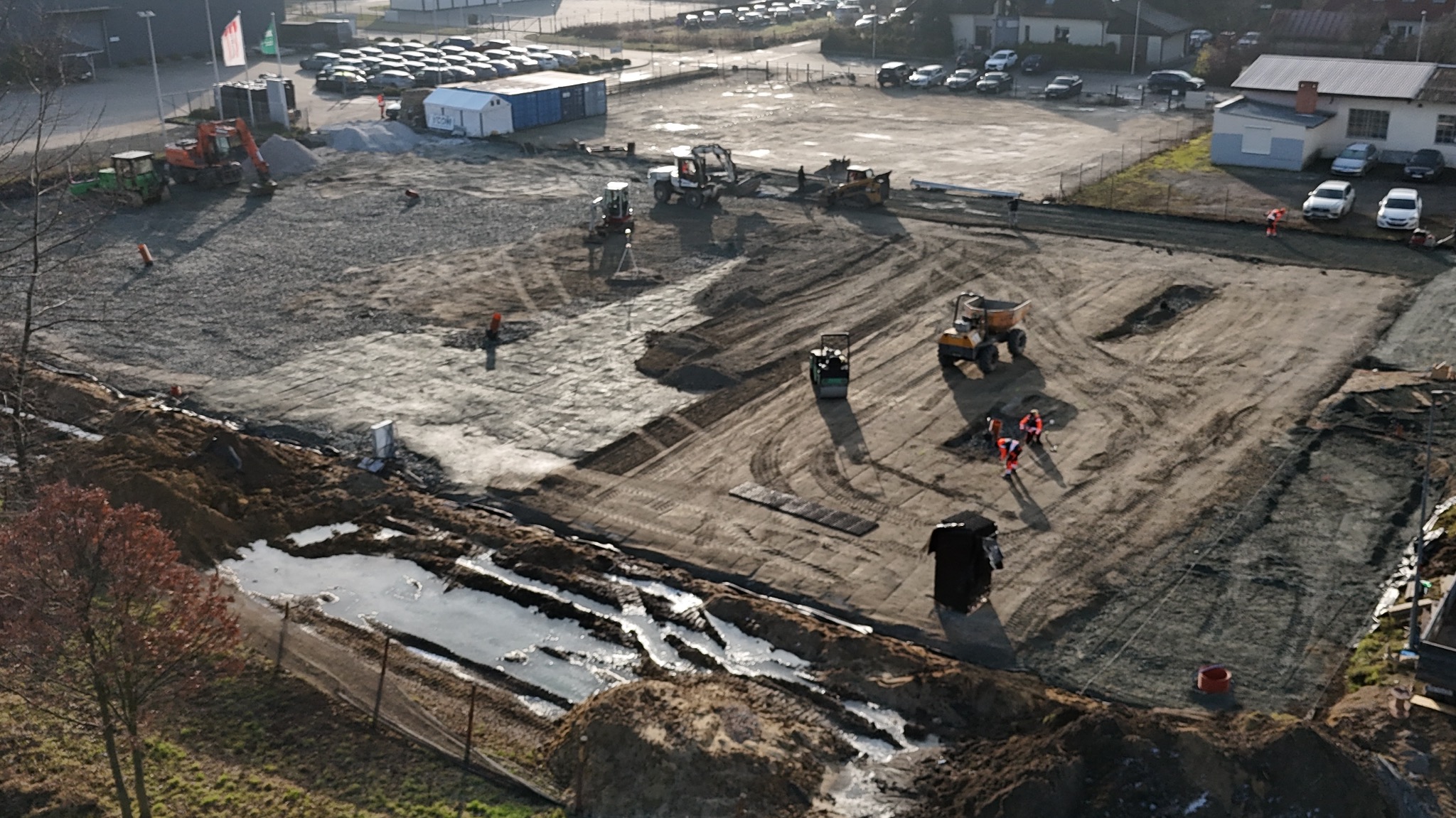 Z góry widok placu budowy z ciężkim sprzętem, w tym walcami i koparkami, przygotowującego teren pod budowę, z widocznymi śladami opon i pracownikami w pomarańczowych kombinezonach.