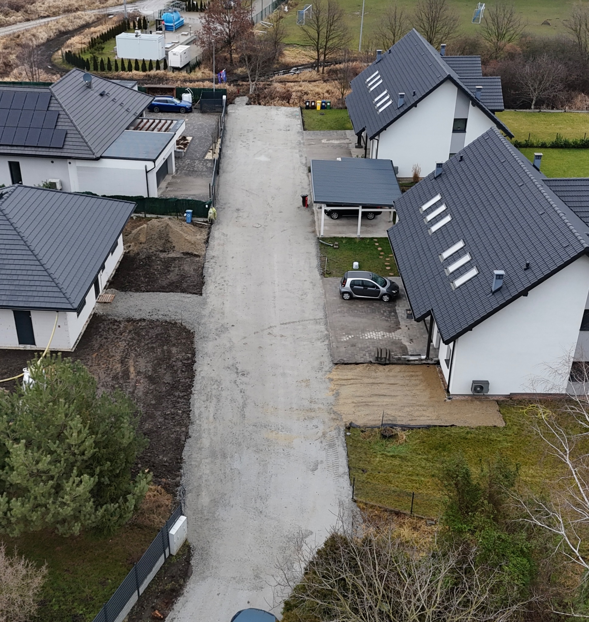 Widok z góry na teren budowy z nowymi domami jednorodzinnymi, gdzie droga wewnętrzna jest w trakcie przygotowania, z widocznym materiałem sypkim i lekkim sprzętem budowlanym.