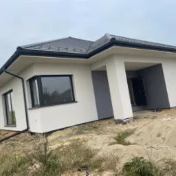 Dom jednorodzinny z nową, jasną elewacją i ciemnoszarą stolarką okienną, otoczony niezagospodarowanym terenem z widocznymi śladami prac budowlanych, w tle pochmurne niebo.