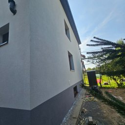 SOL-BUD - Dwukolorowa elewacja domu jednorodzinnego z widocznymi pracownikami na rusztowaniu, fragmentem iglastego drzewa w kadrze i resztkami styropianu na ziemi.