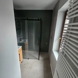 Nowoczesna łazienka z kabiną prysznicową typu walk-in z czarnymi ramami, umywalką z drewnianą szafką, zielonymi heksagonalnymi płytkami na ścianie i grzejnikiem drabinkowym przy oknie na poddaszu.