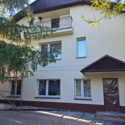 SOL-BUD - Dwupiętrowy dom z balkonem po renowacji elewacji, z brązowym dachem i kremową fasadą. Wejście zabezpieczone folią, widoczne rusztowanie i kostka brukowa.