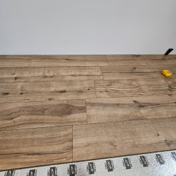 SOL-BUD - Układanie paneli podłogowych na podkładzie izolacyjnym. Widoczna żółta miarka i fragment ściany. Jasne panele imitujące drewno, ułożone w regularny wzór.