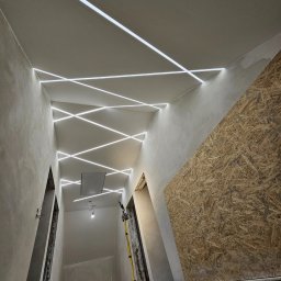 SOL-BUD - Wnętrze w trakcie wykończenia z oryginalnym oświetleniem sufitowym LED w geometryczne wzory. Ściany surowe, częściowo zakryte płytą OSB. Widoczny statyw oświetleniowy.