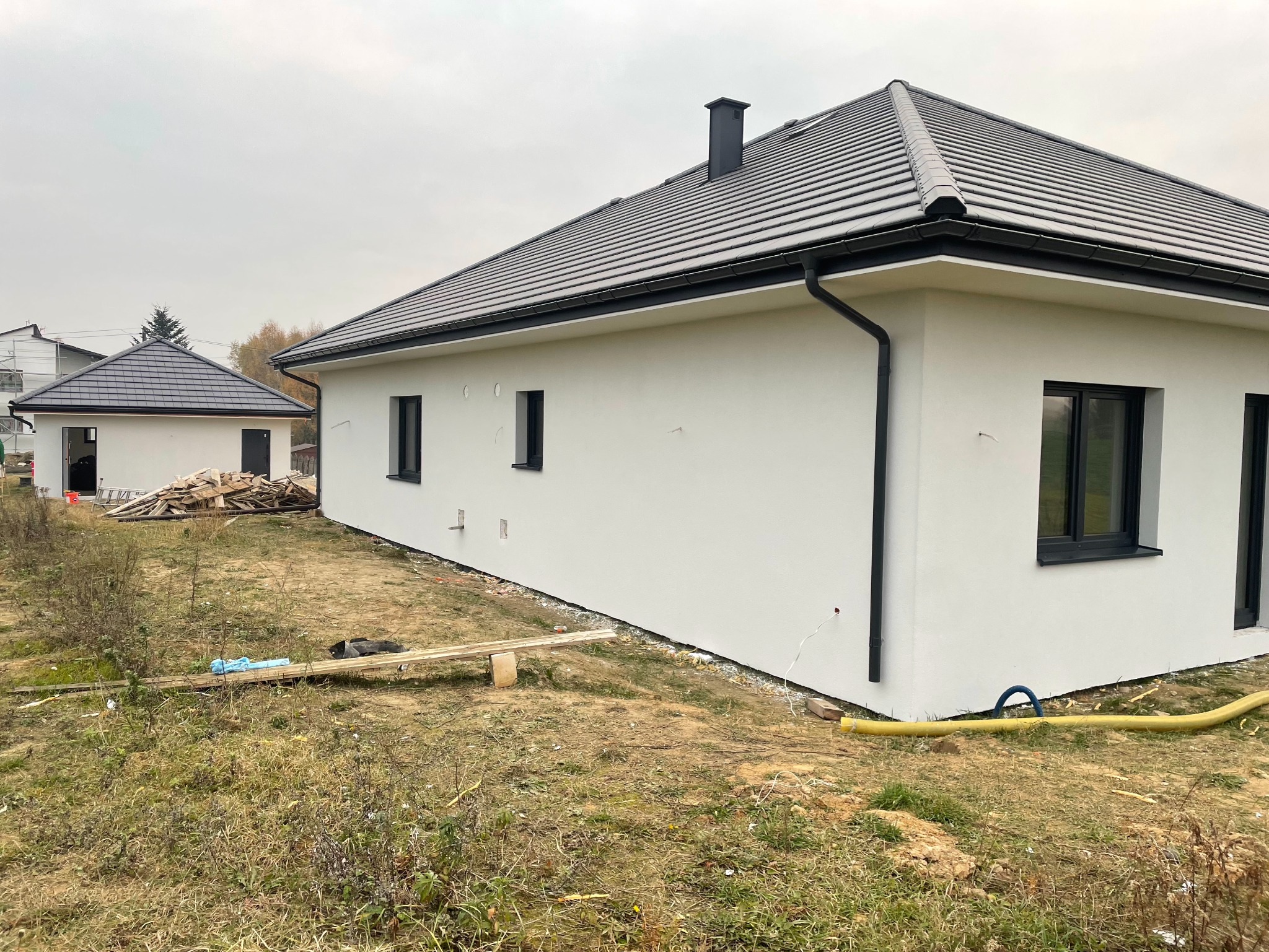 Nowoczesny dom jednorodzinny z białą elewacją i grafitową dachówką, widok z boku, z fragmentem drugiego budynku w tle, teren wokół w stanie surowym.
