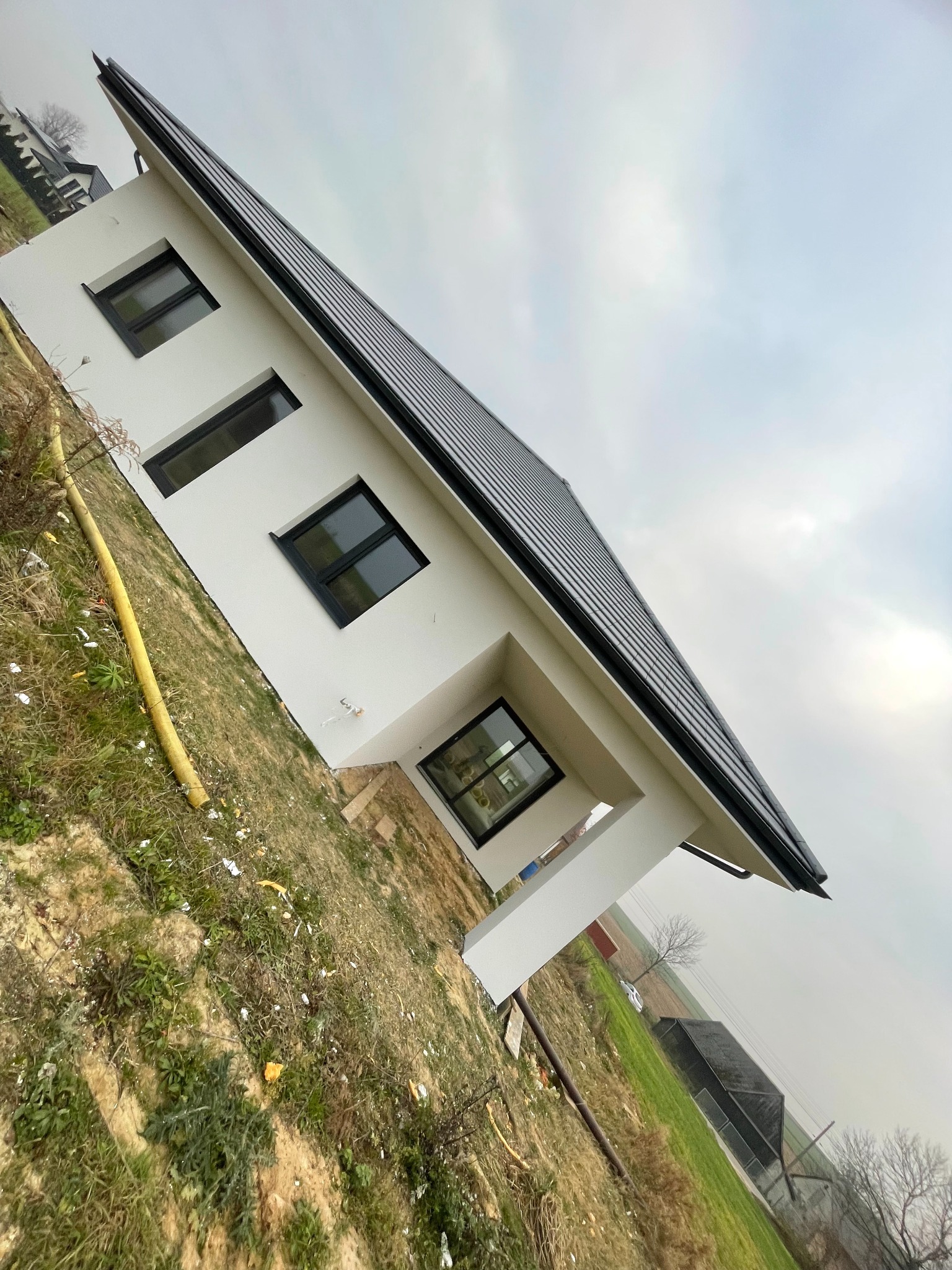 Nowoczesny dom jednorodzinny z czarną dachówką i białymi ścianami zewnętrznymi w Trzyciąż, uchwycony pod kątem, z widoczną żółtą rurą na trawniku i pochmurnym niebem w tle.