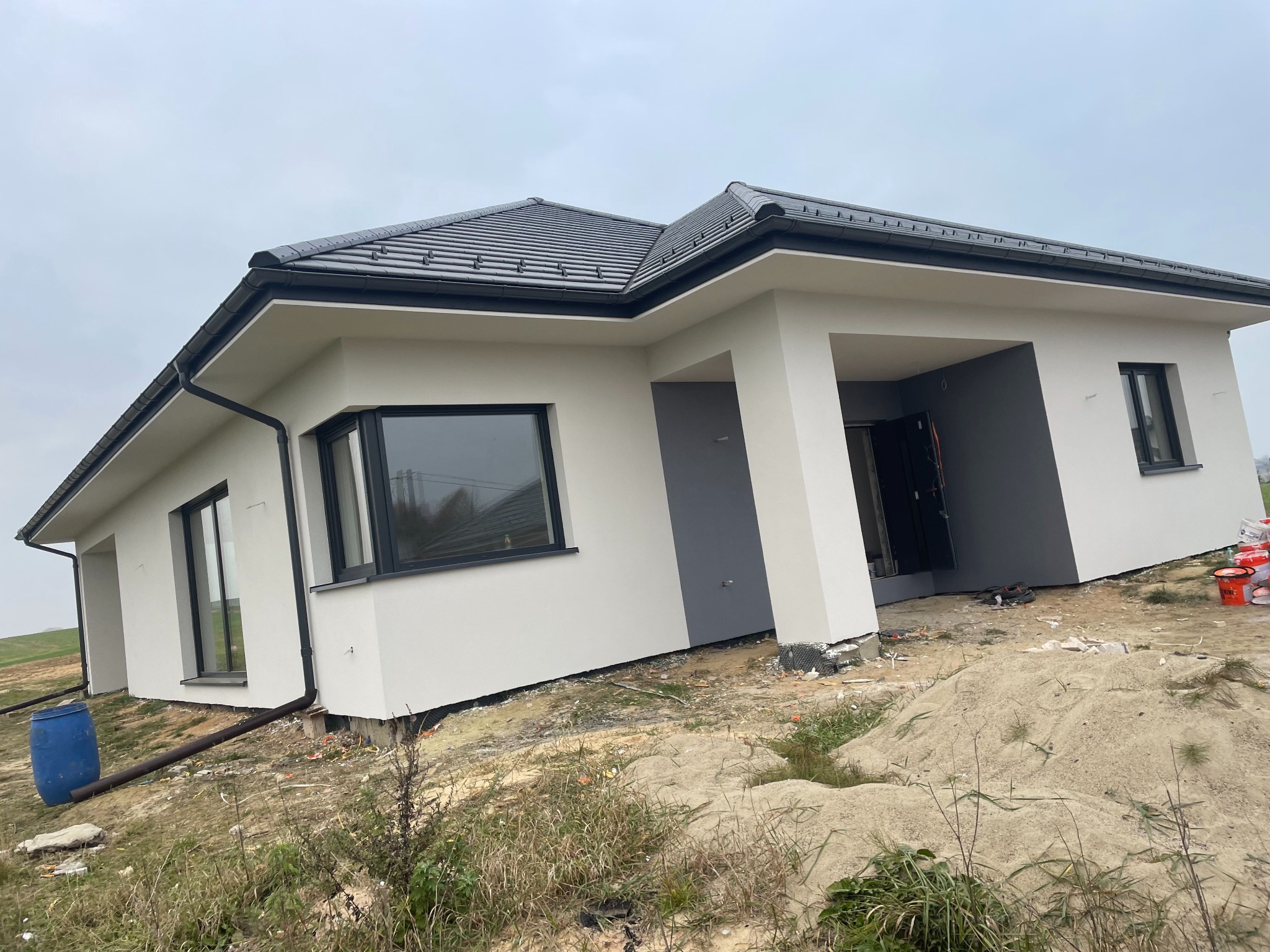 Dom jednorodzinny z nową, jasną elewacją i ciemnoszarą stolarką okienną, otoczony niezagospodarowanym terenem z widocznymi śladami prac budowlanych, w tle pochmurne niebo.