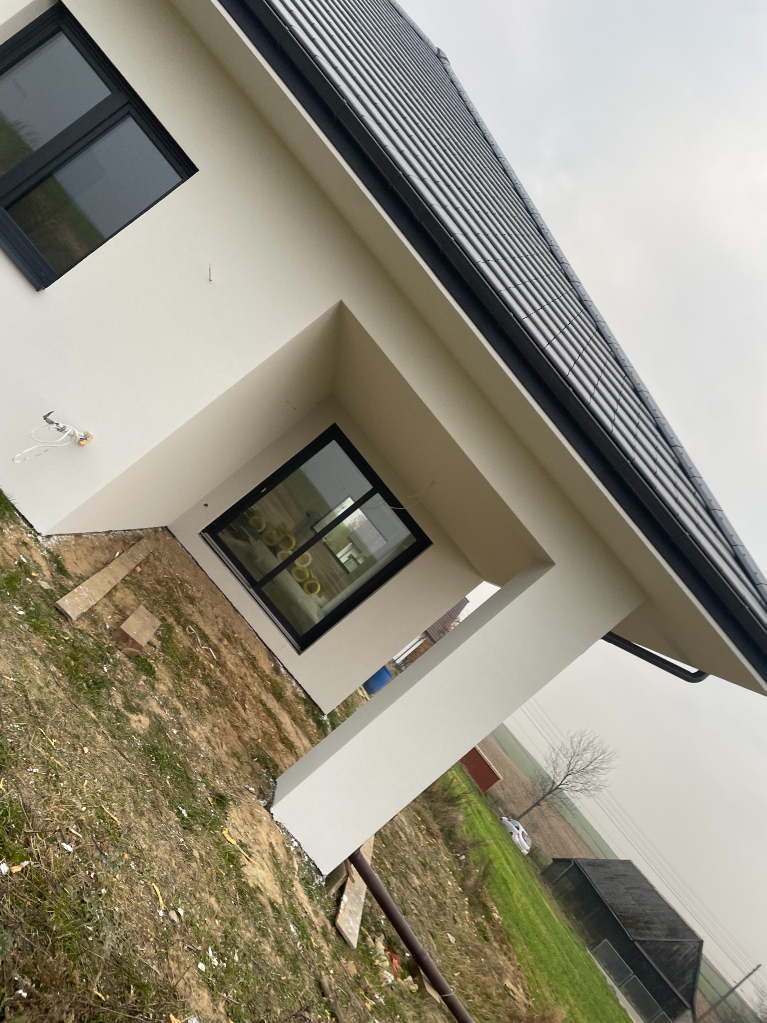 Fragment nowo wybudowanego domu z białymi ścianami, czarnymi oknami i grafitowym dachem, widok z dołu, na zewnątrz widoczne pole i drzewo w oddali.
