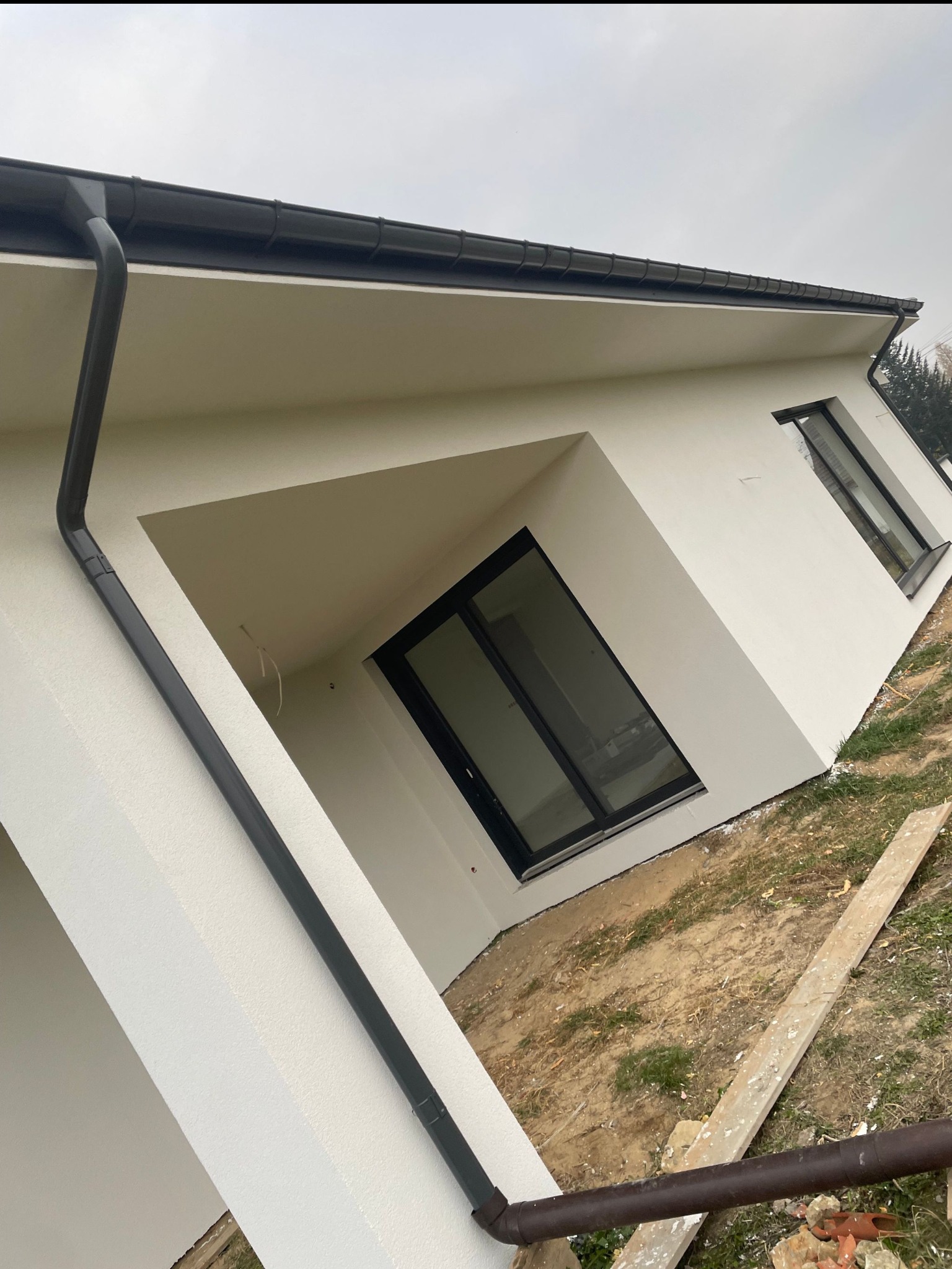 Nowoczesny dom jednorodzinny z jasną elewacją, czarnymi oknami i orynnowaniem, widok z dołu, teren wokół częściowo zagospodarowany, z fragmentami trawy i gruzu.