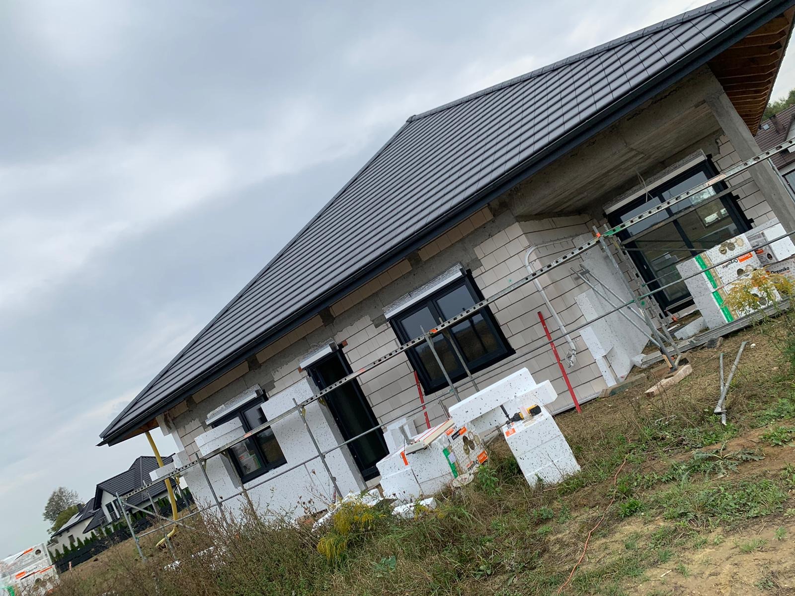 Dom jednorodzinny w trakcie prac, widoczne ocieplenie ścian styropianem, czarne okna, szare dachówki, rusztowania, ziemia z trawą, pochmurne niebo.