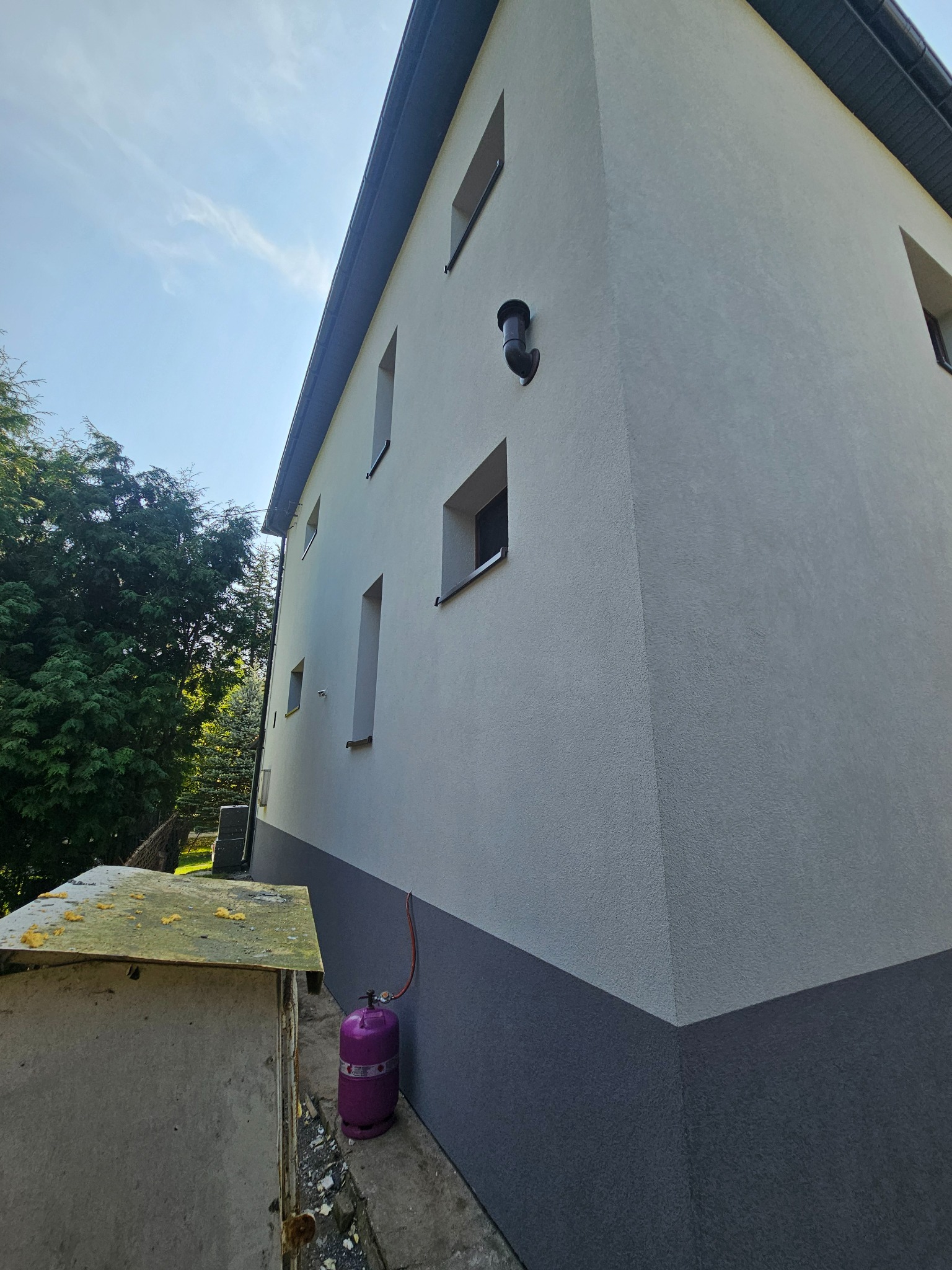 Dwukondygnacyjny budynek z jasną elewacją i ciemnym cokołem, widoczny z dołu pod kątem, z butlą gazową przy ścianie i fragmentem betonowej konstrukcji w kadrze.