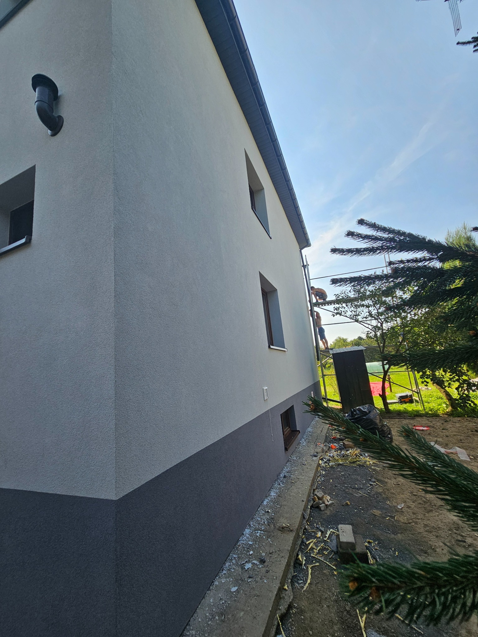 Dwukolorowa elewacja domu jednorodzinnego z widocznymi pracownikami na rusztowaniu, fragmentem iglastego drzewa w kadrze i resztkami styropianu na ziemi.