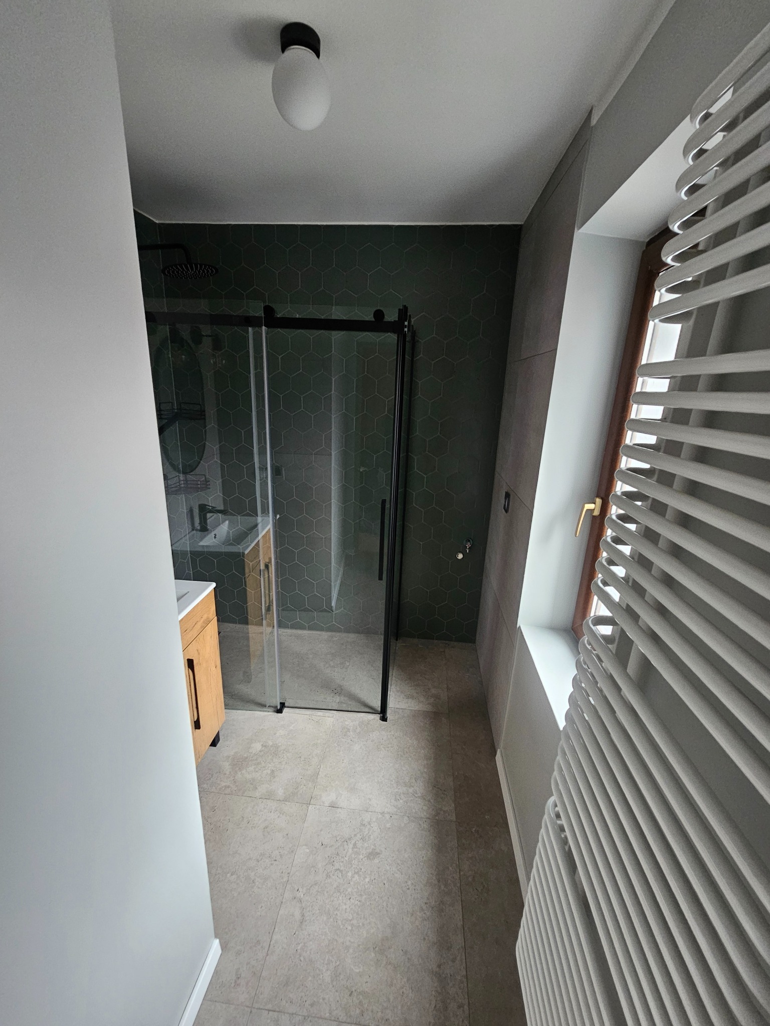 Nowoczesna łazienka z kabiną prysznicową typu walk-in z czarnymi ramami, umywalką z drewnianą szafką, zielonymi heksagonalnymi płytkami na ścianie i grzejnikiem drabinkowym przy oknie na poddaszu.