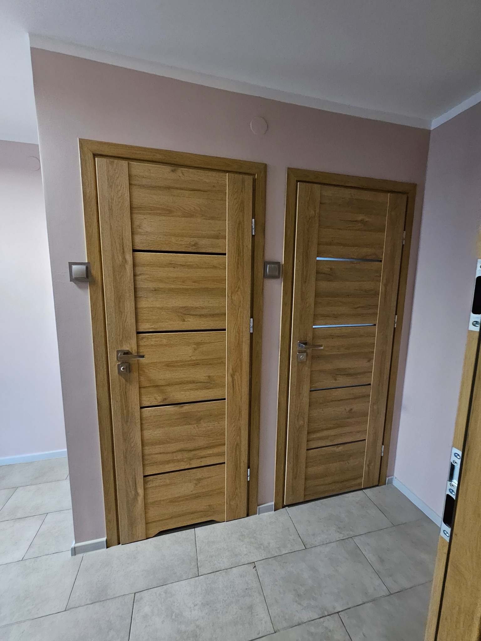 Dwa drewniane drzwi wewnętrzne z poziomymi panelami, zamontowane w korytarzu z płytkami podłogowymi i ścianami pomalowanymi na różowo-beżowy odcień.