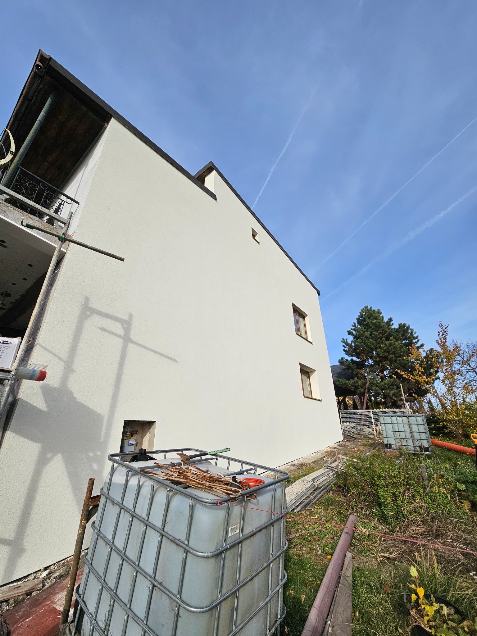 Biała elewacja domu jednorodzinnego po tynkowaniu, widok z dołu, z elementami rusztowania i zbiornikiem na wodę w kadrze. Błękitne niebo w tle.