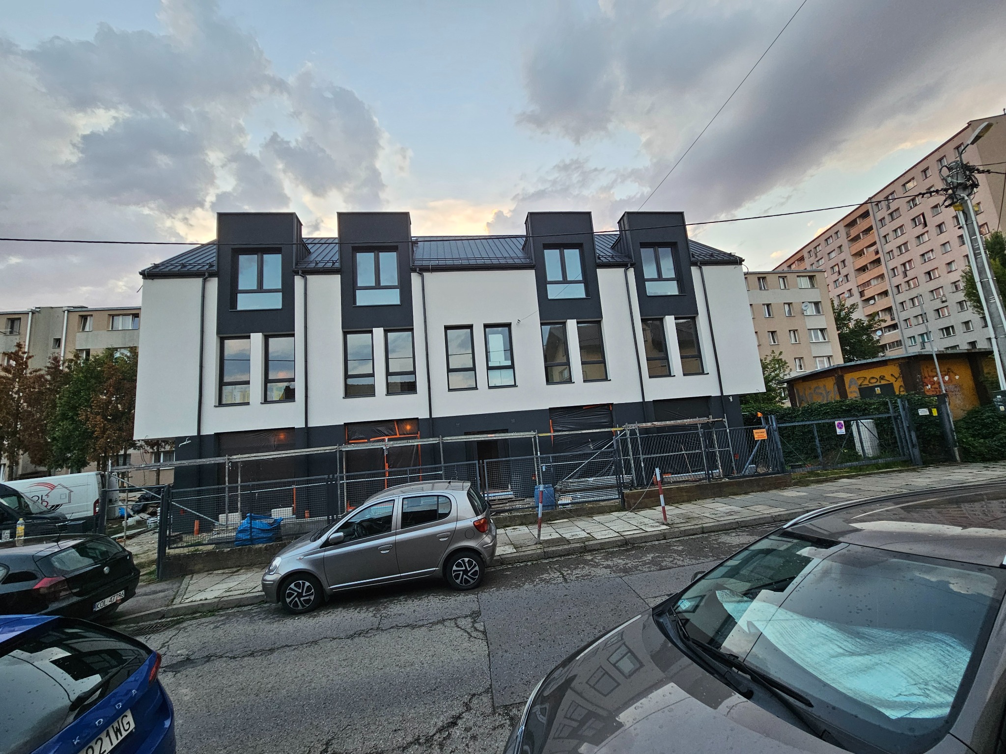 Nowoczesny budynek z białą elewacją i czarnymi akcentami, w trakcie wykończenia. Widoczne rusztowania i zaparkowane samochody. Pochmurne niebo w tle.