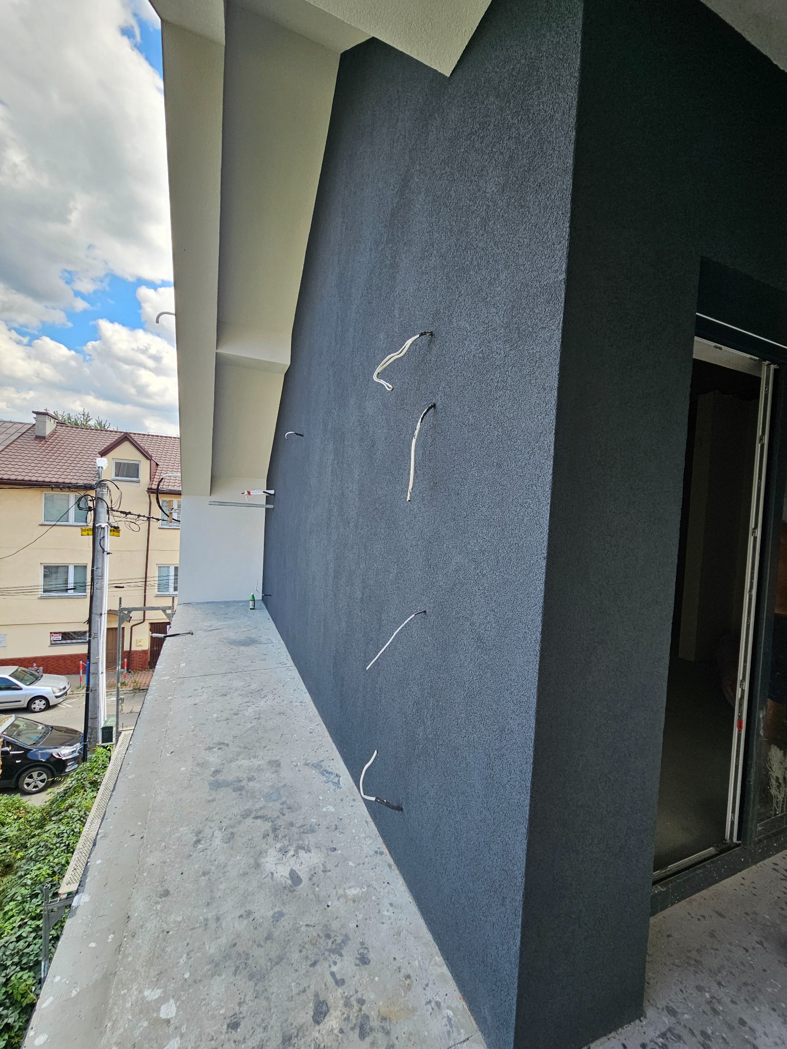 Szara, ziarnista elewacja budynku z wystającymi elementami montażowymi, widok z balkonu na okolicę i fragment sąsiedniego budynku. Nowoczesne wykończenie.