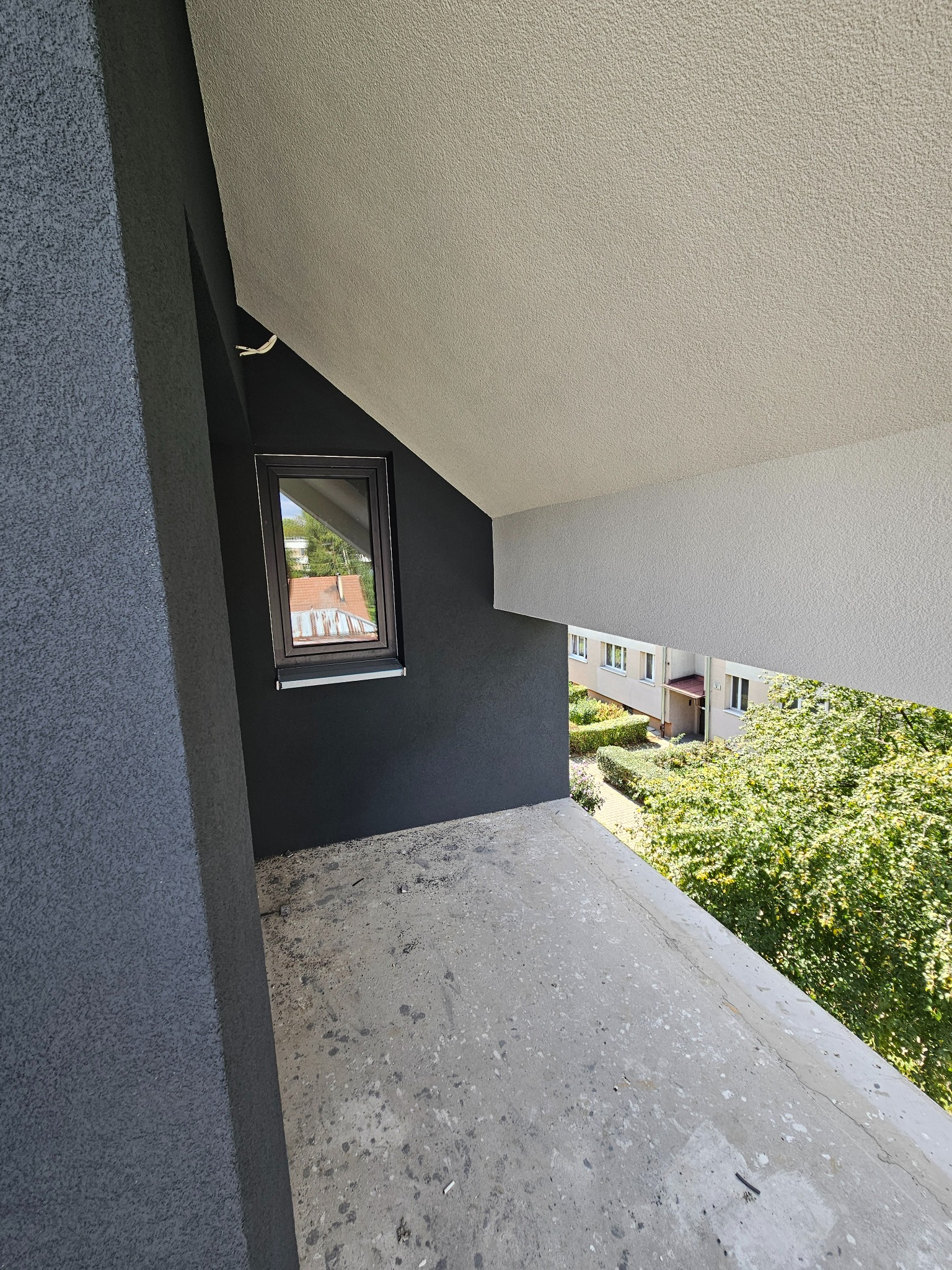 Szary balkon z widokiem na zieleń, czarna ściana z oknem. Surowy beton na podłodze kontrastuje z odnowioną elewacją. Widok z góry.