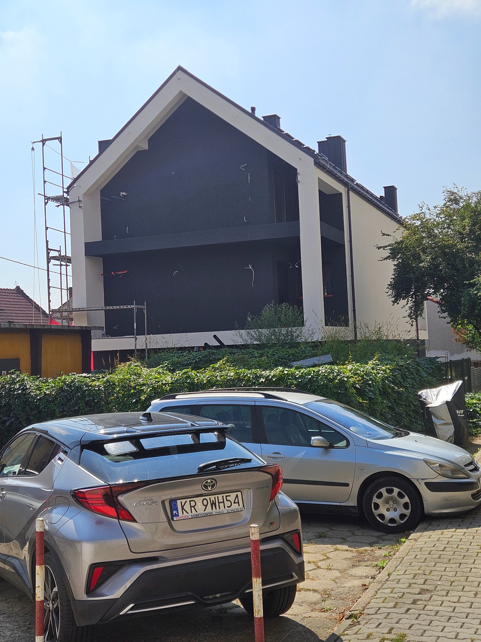 Nowoczesny dom z czarną elewacją w trakcie wykończenia, widoczne rusztowania. Na pierwszym planie zaparkowane samochody i zieleń.