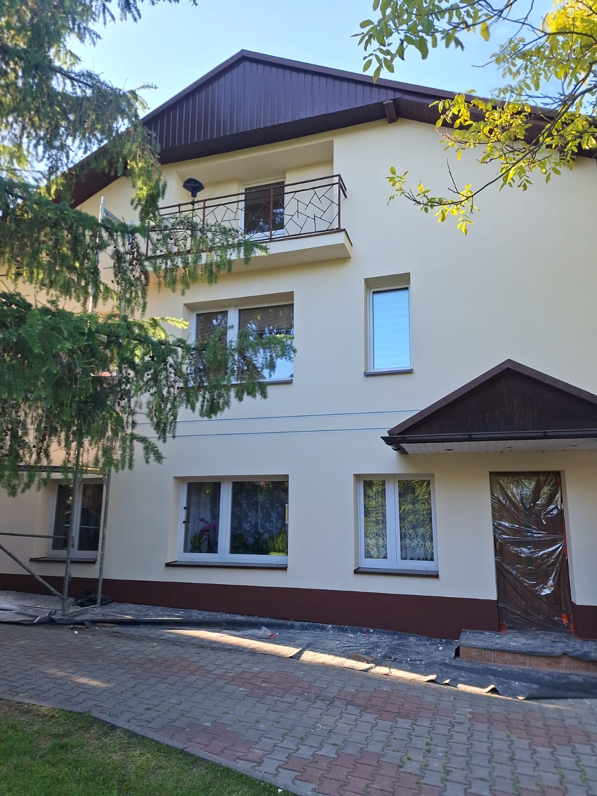 Dwupiętrowy dom z balkonem po renowacji elewacji, z brązowym dachem i kremową fasadą. Wejście zabezpieczone folią, widoczne rusztowanie i kostka brukowa.