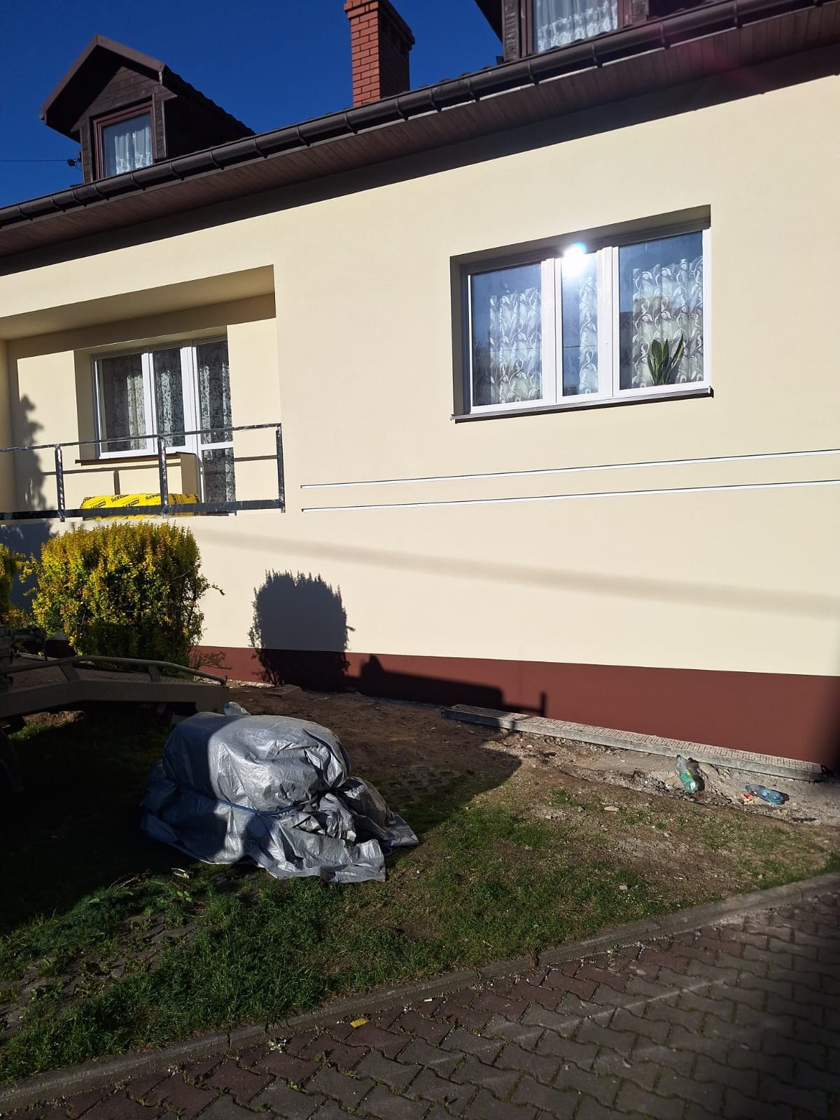 Jasna elewacja domu jednorodzinnego po renowacji, z widocznymi detalami wykończenia i elementami przygotowania terenu wokół budynku.