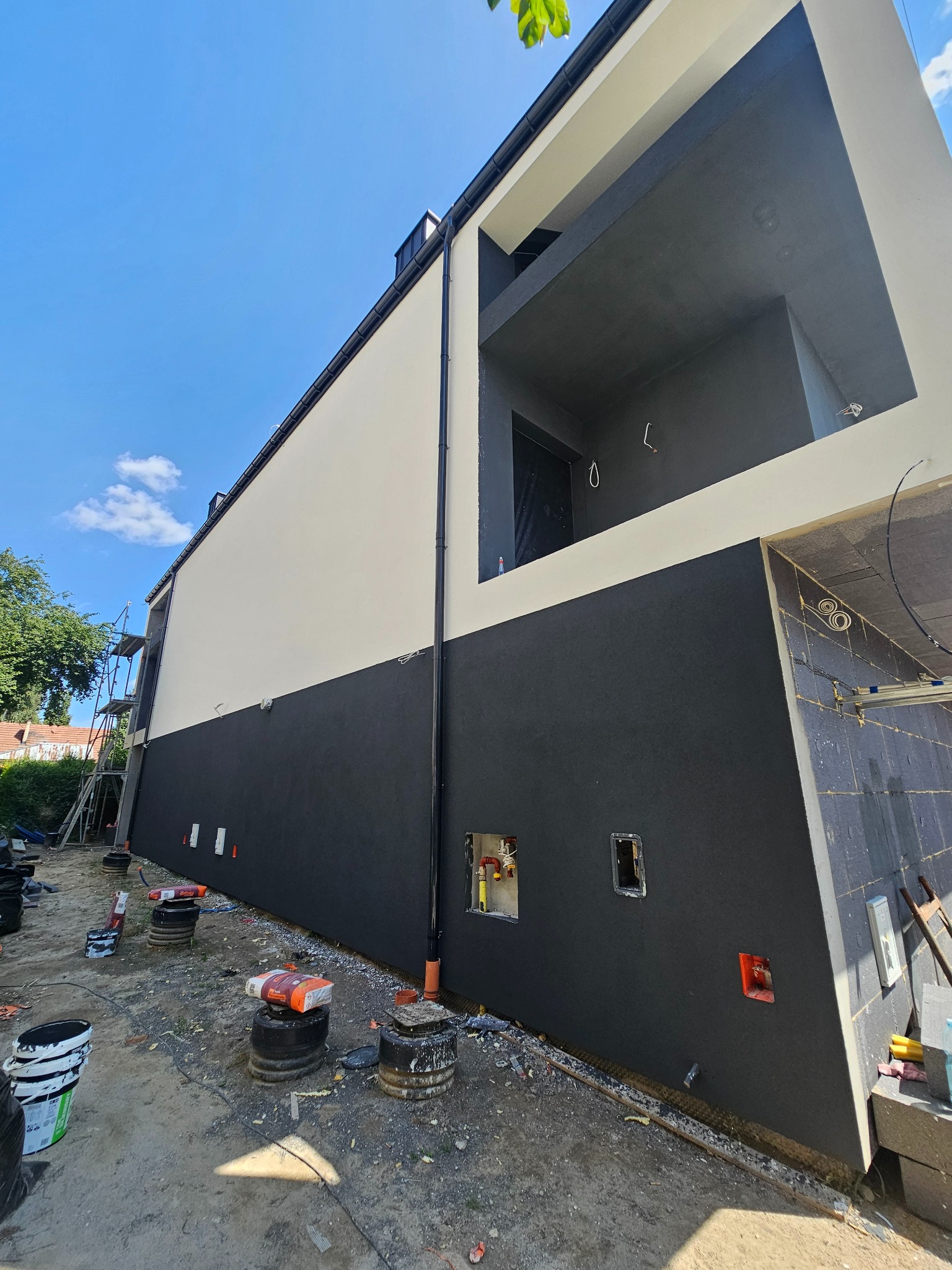 Dwukolorowa elewacja budynku w trakcie wykończenia, z widocznymi elementami instalacji i materiałami budowlanymi na placu budowy. Ujęcie z dołu, błękitne niebo w tle.