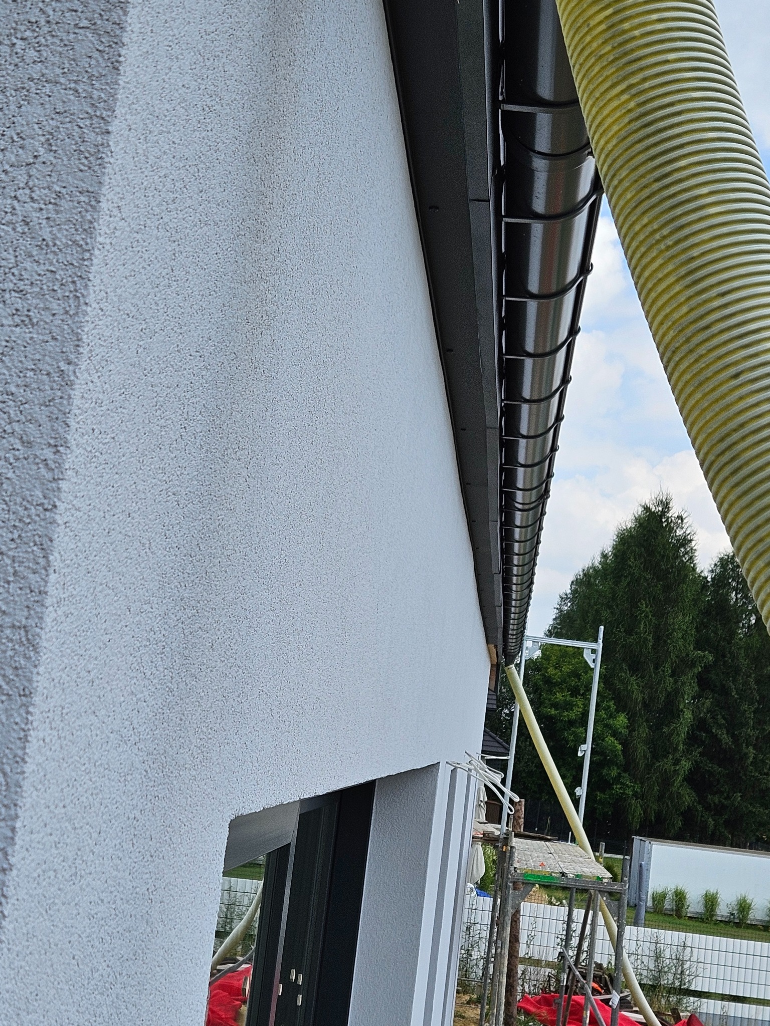 Szara elewacja budynku z widocznym systemem rynnowym i żółtym przewodem wentylacyjnym. W tle zieleń drzew i fragment rusztowania.