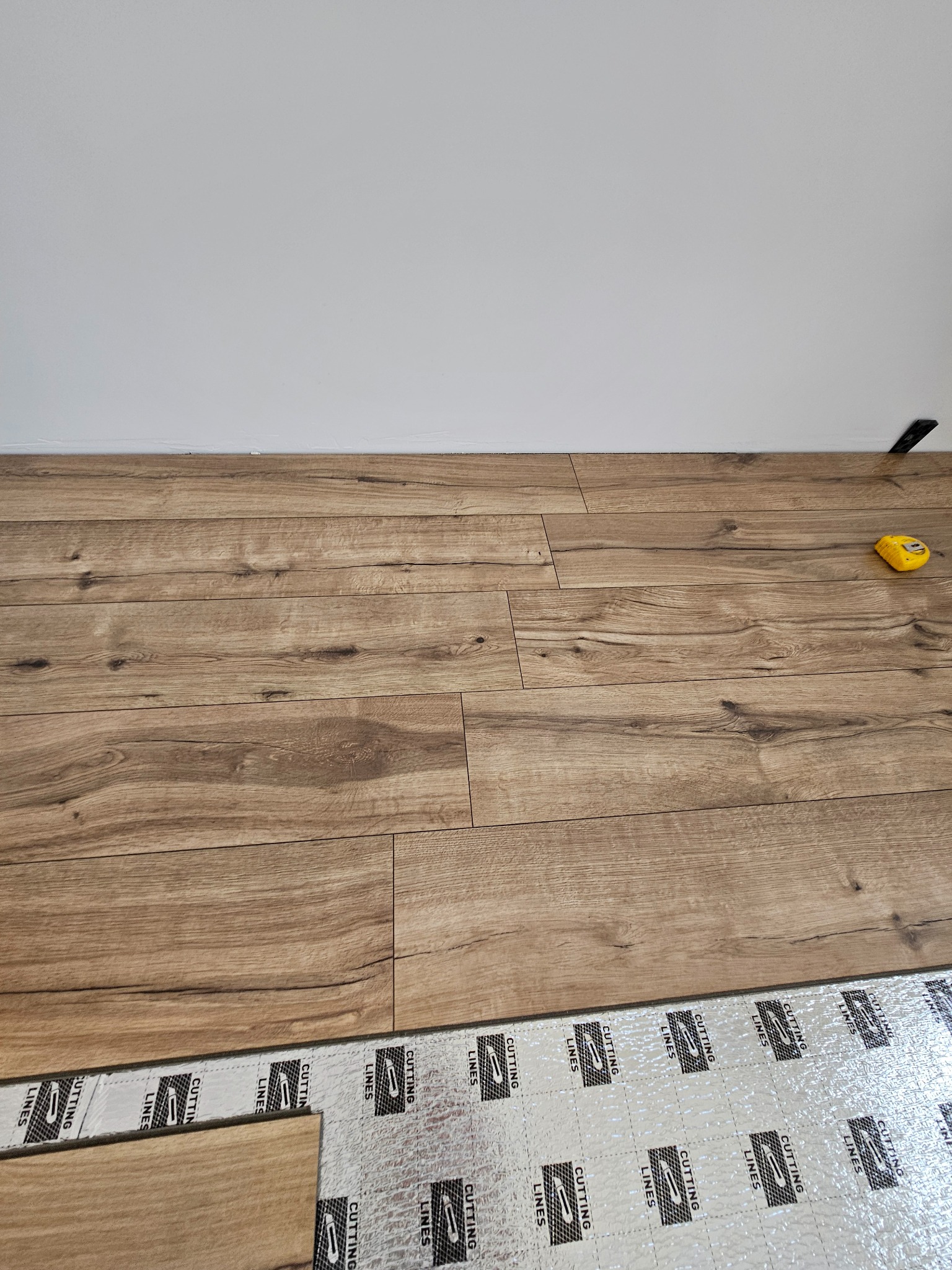 Układanie paneli podłogowych na podkładzie izolacyjnym. Widoczna żółta miarka i fragment ściany. Jasne panele imitujące drewno, ułożone w regularny wzór.