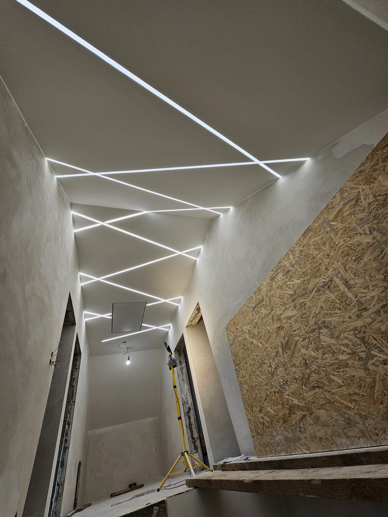 Wnętrze w trakcie wykończenia z oryginalnym oświetleniem sufitowym LED w geometryczne wzory. Ściany surowe, częściowo zakryte płytą OSB. Widoczny statyw oświetleniowy.