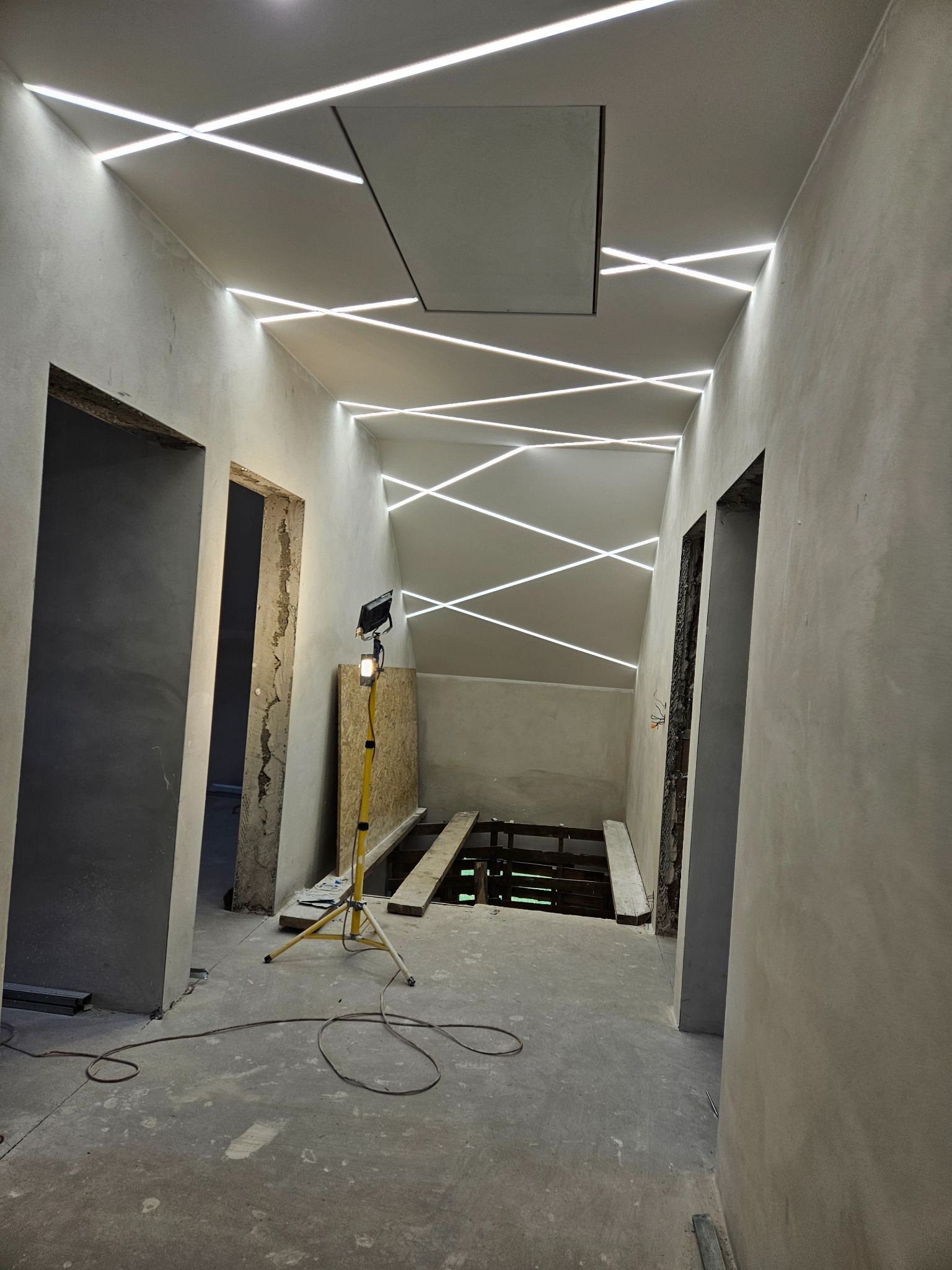 Korytarz w trakcie wykończenia z oryginalnym oświetleniem sufitowym LED w kształcie krzyżujących się linii. Widoczne surowe ściany i podłoga, lampa robocza.