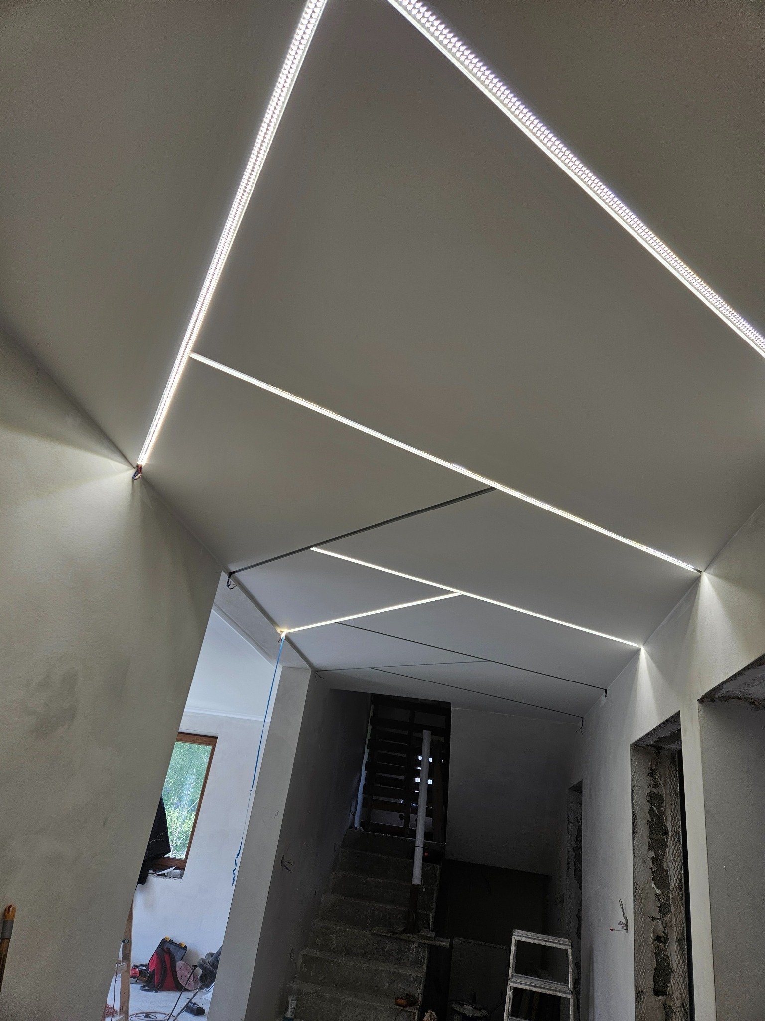 Nowoczesny sufit podwieszany z geometrycznym układem oświetlenia LED w trakcie montażu. Widoczne niedokończone ściany i schody.