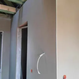 Świeżo nałożony tynk maszynowy na ścianie wewnętrznej, widoczne wystające kable i puszki elektryczne, surowe drewniane elementy konstrukcji stropu.