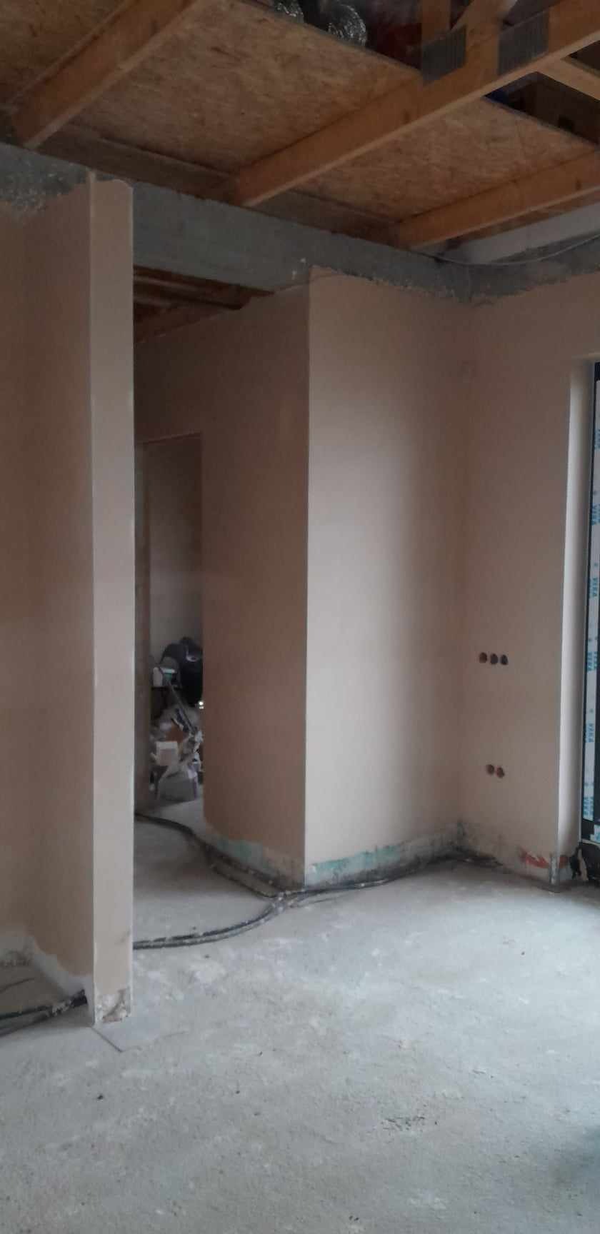 Wnętrze budynku w trakcie prac wykończeniowych: ściany pokryte świeżym, jasnym tynkiem, widoczne puszki elektryczne bez osprzętu, surowy beton na podłodze, drewniana konstrukcja stropu.