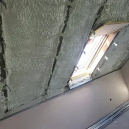 Poddasze z oknem dachowym po aplikacji pianki izolacyjnej, widoczne elementy konstrukcyjne i profile montażowe, jasne światło dzienne.