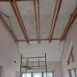 Poddasze z drewnianą konstrukcją pokryte jednolitą warstwą białej pianki izolacyjnej, widok z dołu, z metalowym rusztowaniem w pomieszczeniu.