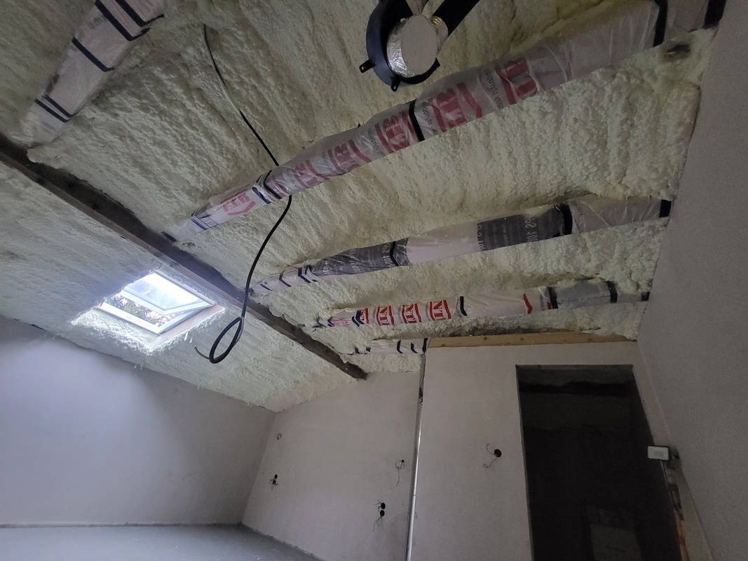 Poddasze po aplikacji pianki PUR, widoczne zabezpieczone rury wentylacyjne i okno dachowe. Ściany częściowo otynkowane, przygotowane pod wykończenie.