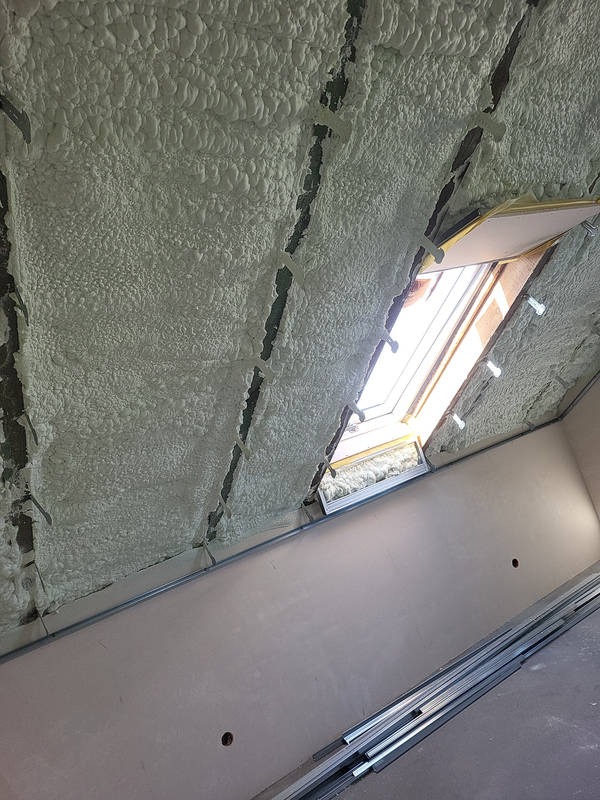 Poddasze z oknem dachowym po aplikacji pianki izolacyjnej, widoczne elementy konstrukcyjne i profile montażowe, jasne światło dzienne.