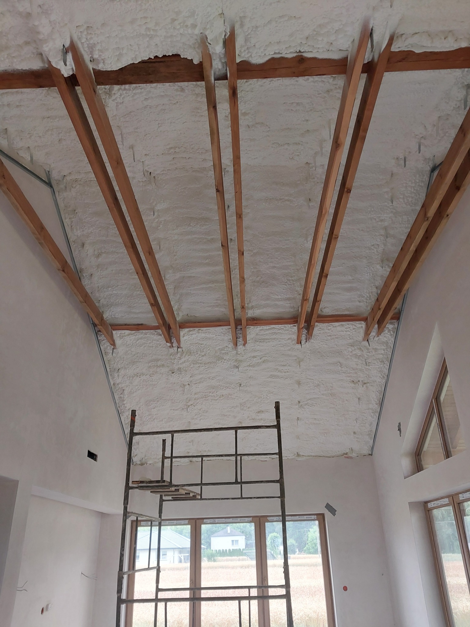 Poddasze z drewnianą konstrukcją pokryte jednolitą warstwą białej pianki izolacyjnej, widok z dołu, z metalowym rusztowaniem w pomieszczeniu.