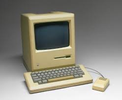 Komputer Macintosh 128K z 1984 roku, z oddzielną klawiaturą i myszą, na jednolitym szarym tle. Widoczne ślady użytkowania na obudowie.