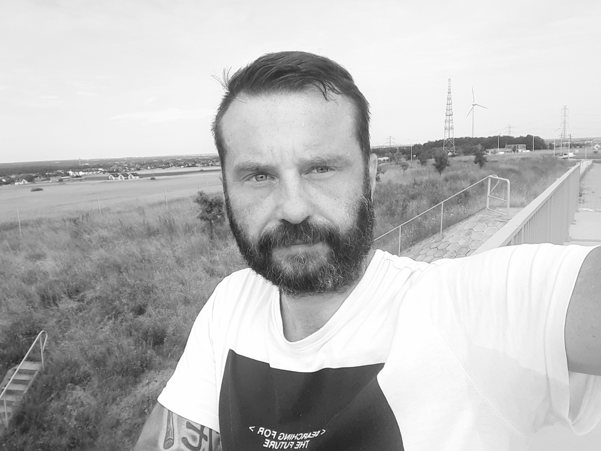 Mężczyzna z brodą robiący selfie na tle krajobrazu z wiatrakami, ubrany w koszulkę z napisem, widoczny tatuaż na ramieniu.