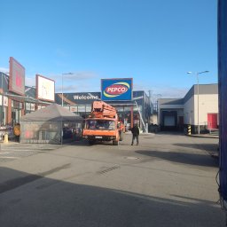 Brak - Pomarańczowy podnośnik koszowy na parkingu przed sklepem Pepco, prawdopodobnie podczas montażu lub demontażu reklamy zewnętrznej.