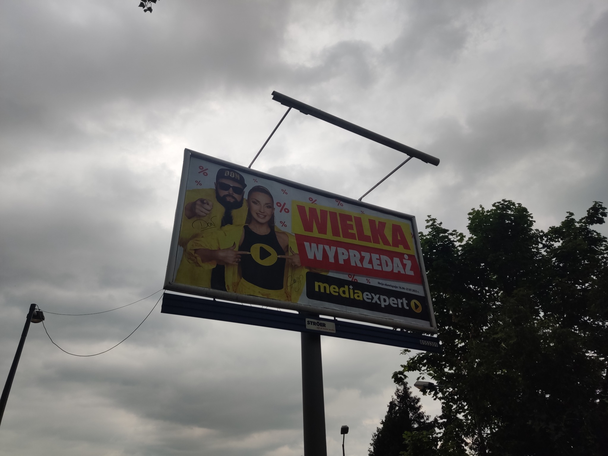 Billboard reklamowy z wizerunkiem celebrytów promujący wyprzedaż w sklepie Media Expert, umieszczony na tle pochmurnego nieba i drzew.