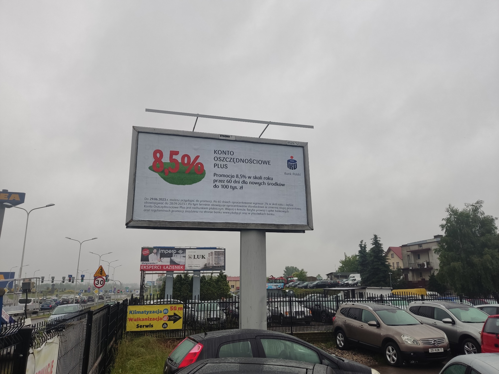 Billboard reklamowy z ofertą konta oszczędnościowego Banku Polski z oprocentowaniem 8,5% na tle pochmurnego nieba i ulicy z samochodami.
