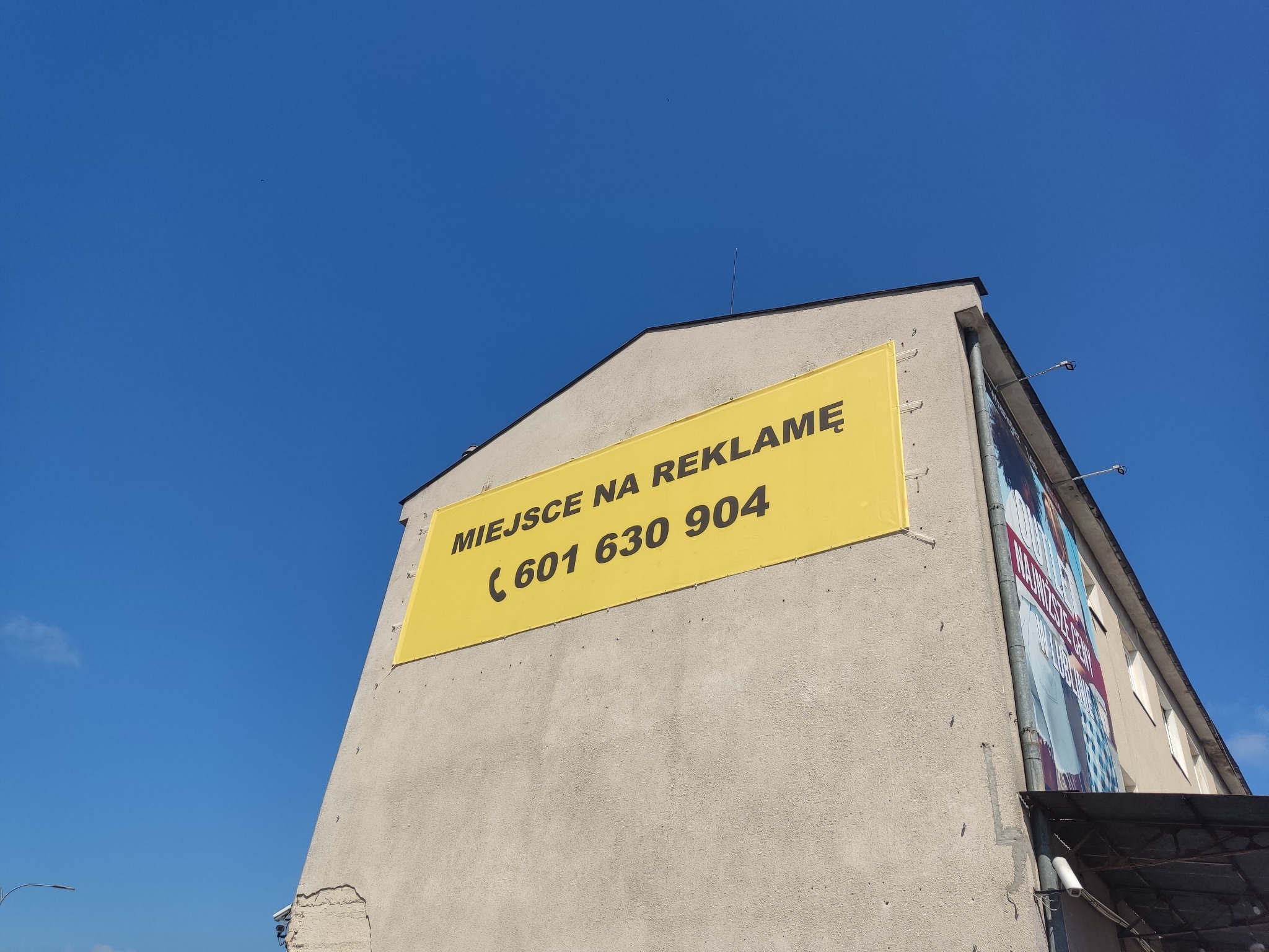 Żółty baner reklamowy z napisem 'Miejsce na reklamę' i numerem telefonu na ścianie budynku, widok z dołu na tle błękitnego nieba.