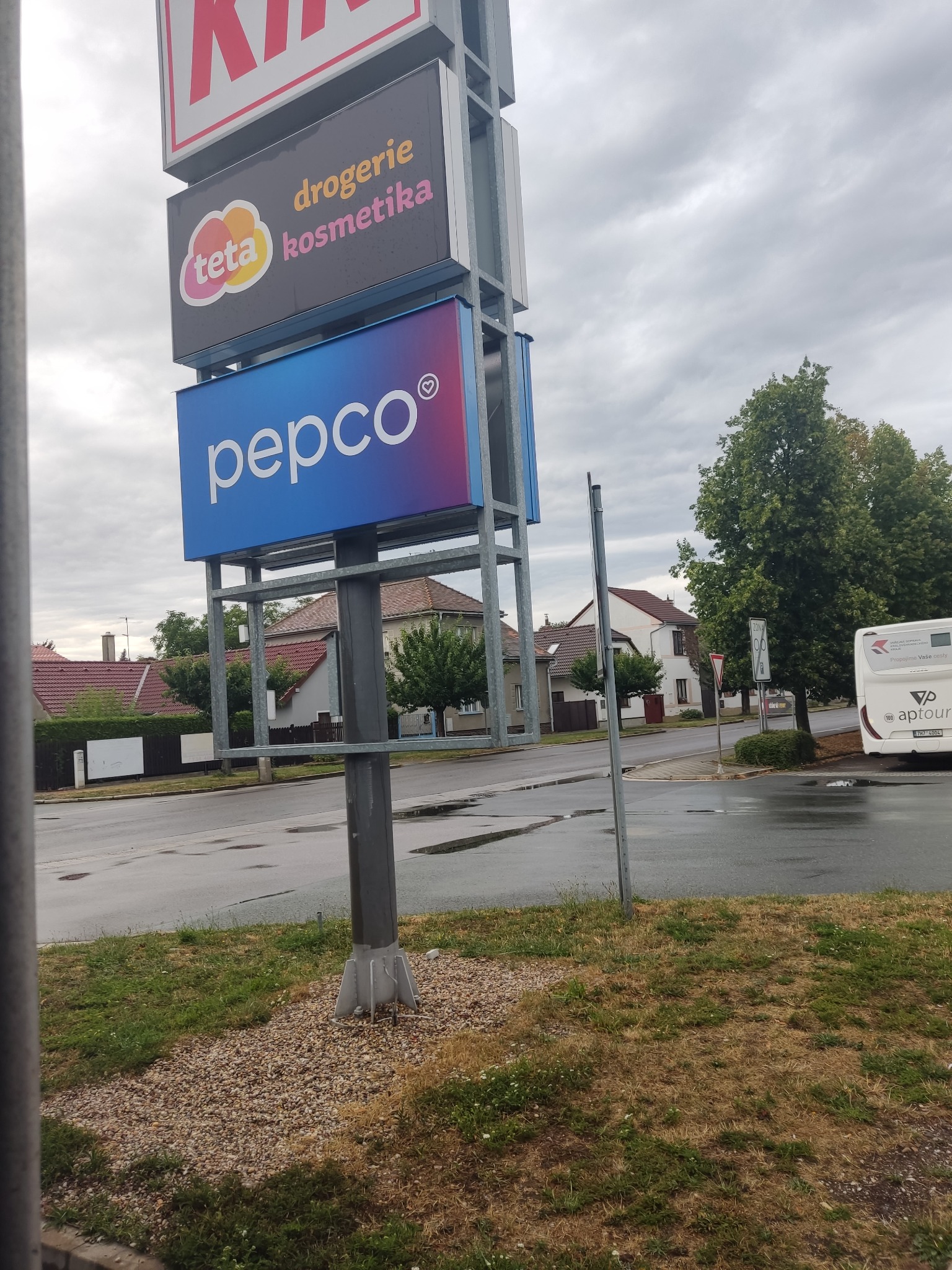 Wysoki pylon reklamowy z logo sieci handlowych na tle ulicy i pochmurnego nieba.