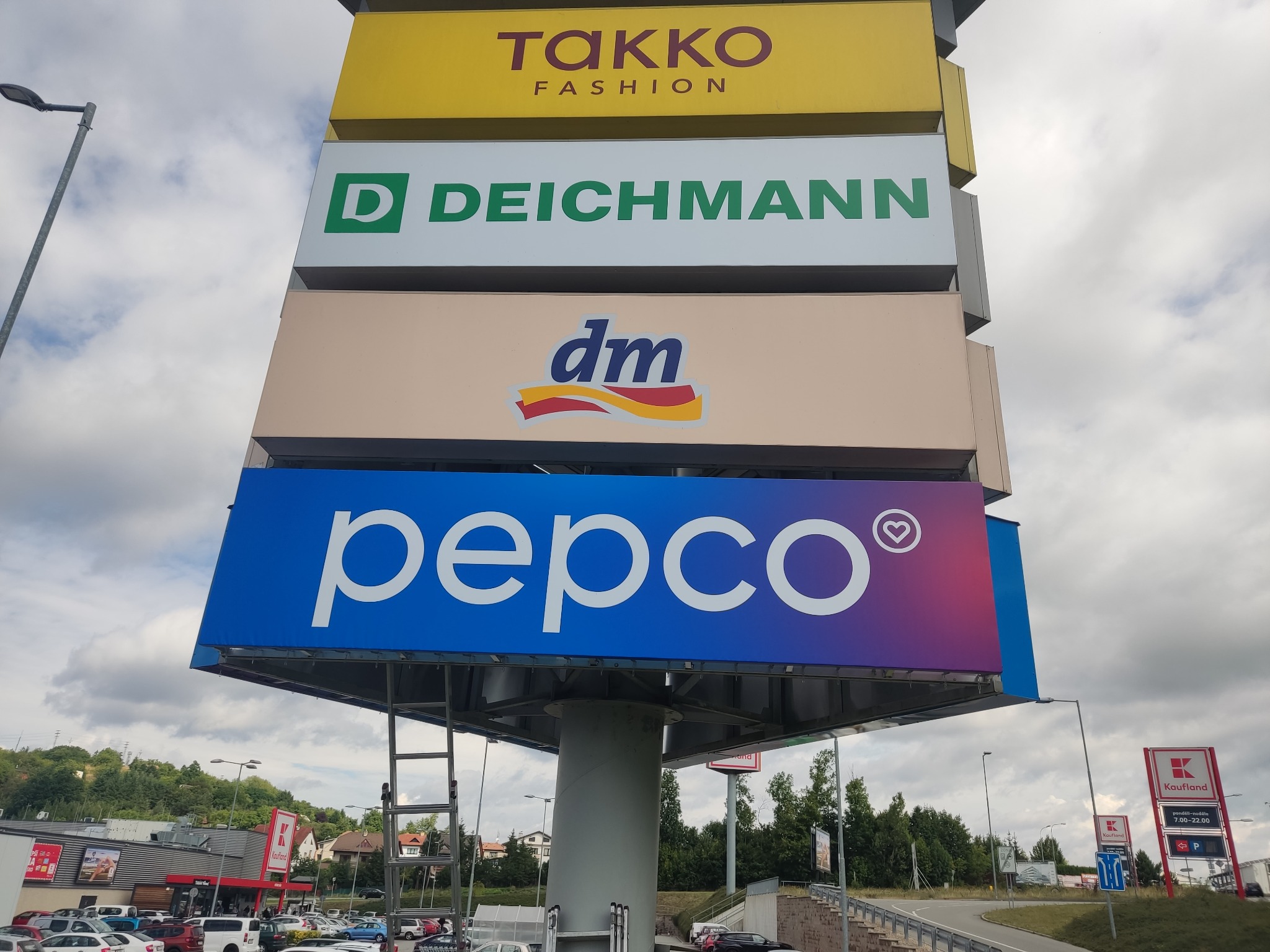Wielopoziomowy pylon reklamowy z logotypami sklepów Takko Fashion, Deichmann, DM i Pepco na tle pochmurnego nieba i parkingu centrum handlowego.