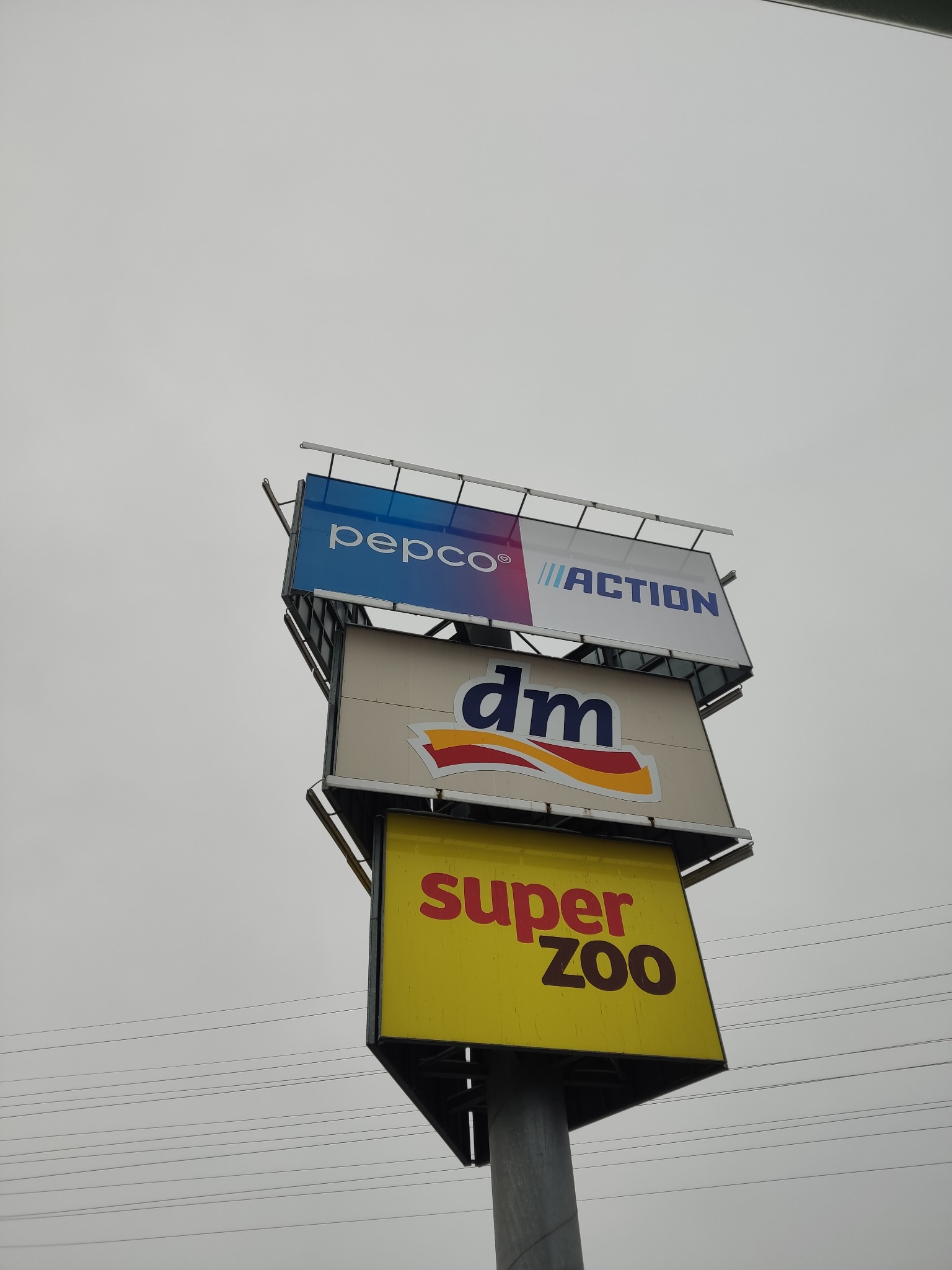 Wielopoziomowy pylon reklamowy z logo Pepco, Action, DM i Super Zoo na tle pochmurnego nieba i linii energetycznych.