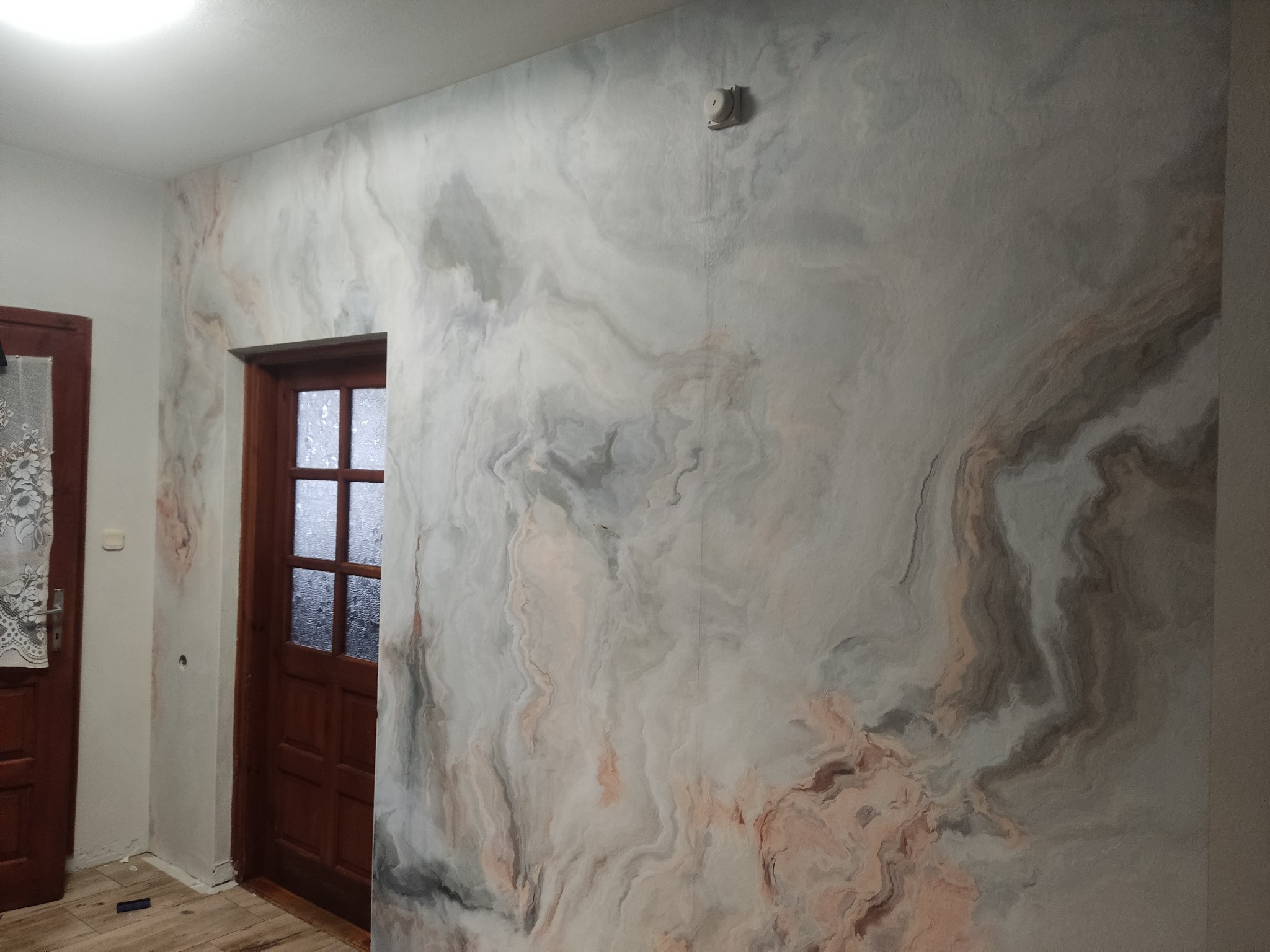 Ściana pokryta tapetą imitującą marmur w odcieniach szarości, bieli i różu, sąsiadująca z drewnianymi drzwiami z szybami.