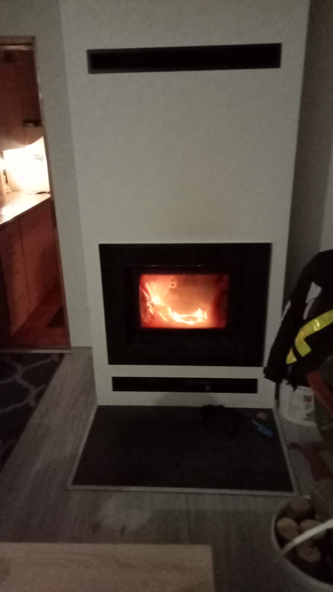 Nowoczesny kominek z białej obudowy z czarną ramą, płomienie widoczne wewnątrz, na podłodze ciemna mata zabezpieczająca.