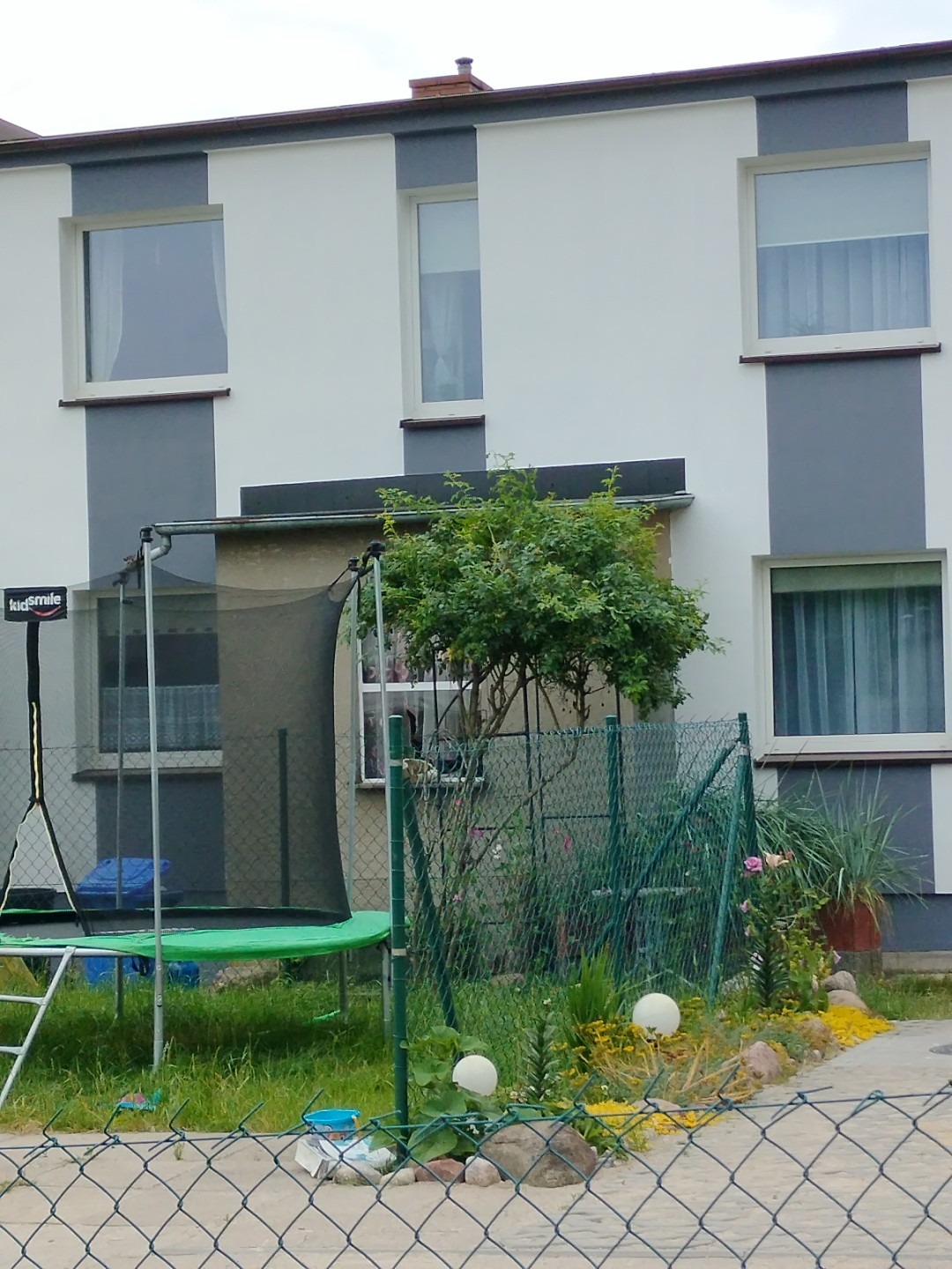 Dwukondygnacyjny dom z szaro-białą elewacją widziany zza zielonego ogrodzenia z siatki. Na pierwszym planie trampolina dla dzieci i dekoracyjne kule ogrodowe.